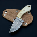 The Texan Knives Custom Handmade Damascus Skinning Knife with a camel bone handle comes with a textured leather sheath and is elegantly displayed on a black background.