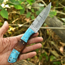 A hand holds the Texan Knives Custom Handmade 5-Inch Small Hunting Knife, featuring a Damascus steel blade and a handle crafted from wood and blue stone with brass rivets, outdoors among green leaves.
