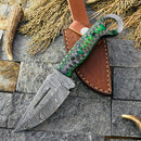The Texan Knives Custom Handmade Small Hunting Knife features a Damascus steel blade and a green and black pakkawood handle, displayed on a brown leather sheath with dried grass and antler pieces nearby.