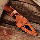 The Texan Knives Custom Handmade Small Hunting Knife features a Damascus steel blade and wooden handle, resting on rustic wood. Its leather sheath with decorative stitching and a buttoned strap highlights the knife's outdoor performance.