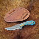 A Texan Knives Winter-Edition Custom Handmade Western Knife with a blue and brown pine cone handle rests beside its decorative brown leather sheath on a furry surface.