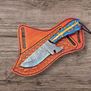 The Texan Knives Custom Handmade Working Cowboy Knife features a Damascus steel blade and a blue-orange pine cone handle, resting on its leather sheath. This unique knife is crafted by Texan Knives.