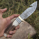 A hand holds the Texan Knives Custom Handmade Damascus Steel Guthook Skinning Knife—9" with stag horn handle, brass guard, and patterned blade. Greenery and a tree trunk appear in the background. Includes leather sheath.