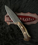 The Texan Knives Custom Forged 1095 High Carbon Steel Hunting Knife features a 12” antler horn handle and comes with a handmade leather sheath, all shown resting on a black fur surface.
