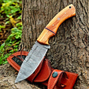 The Texan Knives Custom Handmade 10" Damascus Steel Hunting Knife with Olive Wood Handle & Leather Sheath rests by a tree trunk, the sheath nearby on the ground and green foliage in the background.
