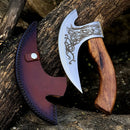 The Texan Knives Custom Hand-Engraved Axe Pizza Cutter, featuring an 8" J2 steel blade and wooden handle, rests against a tree beside its brown leather sheath with black stitching and a snap closure.