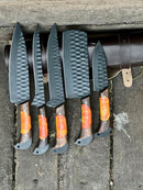 Texan Knives’ Damascus Steel Handmade Chef Knife Set with Sharpener features five knives with textured dark grey blades and orange-accented wooden handles beside a brown leather roll—perfect gift for wife, husband, or Valentine’s Day.