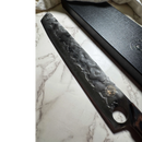 A close-up of the Spaceman Knives Meteor Series Brisket Slicer, featuring a dark patterned blade with a round hole near the handle, rests on white marble beside its black box—precision BBQ carving from Spaceman Knives.