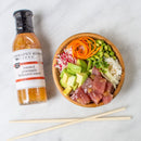 A poke bowl with diced tuna, avocado, carrots, edamame, radish, and rice sits next to a quart bottle of Terrapin Ridge Farms Pineapple Habanero Sauce by Terrapin Ridge Farms. Two chopsticks are placed below the bowl on a white surface.