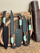 The Texan Knives Premium Knife Set features 13" forged stainless steel blades with elegant wenge wood and resin handles, displayed upright on a round wood slab next to a brown leather sheath.