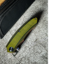 The Spaceman Knives Galatea Folding Pocket Knife, featuring a textured green handle and premium 14C28N steel blade, rests partially open on a grey surface beside a black zippered case—perfect for everyday carry.