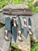 Five Texan Knives “Best Knife Block Set” 13" forged stainless steel chef knives with olivewood and epoxy resin handles, plus leather sheaths, are arranged outdoors on rocks with a grassy background.