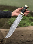 Outdoors near greenery, a person in a dark sweater holds the Texan Knives Authentic Crocodile Dundee Knife—16” handmade Damascus steel blade with brass guard and buffalo horn or leather handle—by a log.