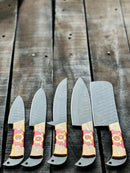 The Texan Knives Stainless Steel Knife Set 13" features five forged blades with olivewood and epoxy handles, arranged on a rustic wooden surface. The varied blades come with a leather sheath, perfect for versatile culinary tasks.
