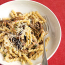 A white bowl of gemelli pasta topped with sautéed mushrooms, Parmesan, and SALTOPIA Infused Sea Salts' Saltopia Truffle Salt, White Italian Truffles: DIRTY WEDDING, sits on a red surface with a fork resting beside it.