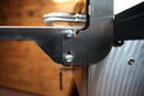 Close-up of a stainless Pimp My Grill® Tactical Stainless Folding Front Shelf for Camp Chef Woodwind 24 / Woodwind Pro 24 / CL 24 attached to a grill with two Phillips head screws.