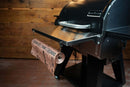 Pimp My Grill Tactical Stainless Folding Front Shelf for Camp Chef Woodwind 24 / Woodwind Pro 24 / CL 24 attached to a pellet grill as a prep workspace, with a roll of branded butcher paper hanging from a hook.