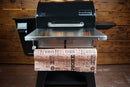 A Pimp My Grill® Tactical Stainless Folding Front Shelf attached to a Camp Chef Woodwind 24 / Woodwind Pro 24 / CL 24, providing a stainless steel workspace with a roll of butcher paper draped over the front.