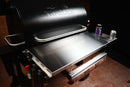 A stainless steel Pimp My Grill Tactical Stainless Folding Front Shelf for Traeger Pro 780 / Ironwood 885 attached to a pellet smoker, featuring an integrated paper roll dispenser underneath and a side mount for drinks and seasonings.