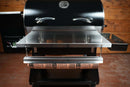 A stainless steel folding front shelf attached to a black pellet grill, featuring a paper dispenser rod holding a roll of butcher paper for this Pimp My Grill® Tactical Stainless Folding Front Shelf for the Traeger Pro 34.