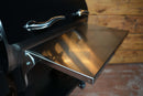 A Pimp My Grill® Tactical Stainless Folding Front Shelf attached to the front of a black Recteq Deck Boss 590 grill.