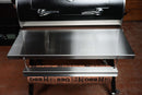 A stainless steel Pimp My Grill® Tactical Stainless Folding Front Shelf attached to a Recteq Patio Legend 410, featuring an integrated paper holder dispensing branded butcher paper to provide convenient grill prep space.