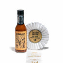 A bottle of California Hot Sauce Solutions CHSS Calexico Rojo Hot Sauce—a Southwest-inspired, fire roasted tomato hot sauce—stands next to a white “Fiery Food Challenge” award ribbon for Hot Sauce, Latin Style – Mild/Medium. Their reflection and “2025 Fiery Food Challenge” text appear below.