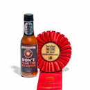 A bottle of California Hot Sauce Solutions Paulman Acre Don’t Pear the Reaper Hot Sauce from California Hot Sauce Solutions stands next to a red Fiery Food Challenge 2025 award ribbon for Reaper Pepper Sauce, both on a white background.