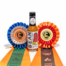A bottle of California Hot Sauce Solutions J's Small Batch BEAN Chili Sauce is displayed between two award ribbons—one blue and one orange—marked “International Flavor Awards,” with attached ribbons reading “2023” and “2023 Spicy.”.