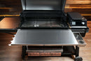 A stainless steel Pimp My Grill® Tactical Stainless Folding Front Shelf attached to a Traeger Ironwood XL, featuring a small storage tray on the right and a roll of butcher paper suspended underneath to maximize grill prep space.