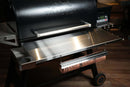 Pimp My Grill® Tactical Stainless Folding Front Shelf attached to a black Traeger Timberline 1300, featuring a side cup holder and a hanging roll of printed butcher paper to expand your outdoor grill workspace.