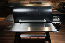 Black pellet grill featuring a Pimp My Grill® Tactical Stainless Folding Front Shelf for the Traeger Timberline 1300, with a paper roll holder attached underneath.