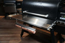 Pimp My Grill® Tactical Stainless Folding Front Shelf for Traeger Timberline 1300 attached to a black pellet grill, featuring a mounted paper roll dispenser underneath as a convenient outdoor grill workspace.