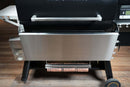 Pimp My Grill® Tactical Stainless Folding Front Shelf attached to a black pellet grill, featuring an integrated roll of butcher paper suspended beneath the stainless steel workspace.
