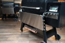 A Pimp My Grill® Tactical Stainless Folding Front Shelf for Traeger Timberline 1300 attached to a black pellet grill, featuring a stainless steel shelf and a paper towel roll holder underneath as an outdoor workspace.