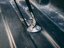 A metal tie-down anchor from the Blue Coolers Secure Universal Tie Down Kit, bolted to a ribbed surface with a black strap attached.
