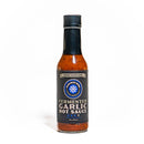 A bottle of California Hot Sauce Solutions Paulman Acre Fermented Garlic Hot Sauce is shown with a black label and cap, highlighted by a blue and white circular logo at the center. This umami-rich hot sauce from California Hot Sauce Solutions displays a reddish-brown color and is set against a white background.