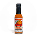 A bottle of California Hot Sauce Solutions Paulman Acre Smoke a Peach Hot Sauce—with a label depicting a red truck hauling an oversized peach—displays the vibrant orange color of this smoked peach hot sauce inside. The label reads "Seasonal Micro Batch," emphasizing its small-batch artistry. Photographed on a white background.