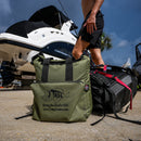 A person in swim trunks walks near a boat, with a Taiga Olive Back Pack Cooler by Taiga Coolers and a black backpack on the ground in the foreground.