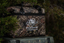 The Taiga Camo soft side 18 pack cooler by Taiga Coolers rests outdoors among greenery—durable camo design, perfect for your next hunting or fishing adventure.