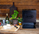 A black pizza oven rests on a wooden countertop beside dough balls, a knife, olive oil, herbs, spices, and potted plants. Nearby is the WPPO Outdoor Cooking Utensil Holder by WPPO LLC Direct, with a wooden cutting board propped against the brown wall in the background.