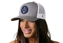 A woman with long brown hair smiles while wearing the Haka Hat Dallas Trucker Hat, a gray and white mesh DAL X Football Snapback by Haka Hat, featuring a round patch with D, A, L letters and a small emblem in the center.