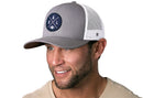 A smiling man wears a Haka Hat Dallas Trucker Hat in gray and white (DAL X Football Snapback) featuring a circular navy logo, paired with a beige T-shirt against a white background.