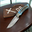 The Texan Knives Premium Damascus Cowboy Knife features a 10" rasp tool steel blade and resin pine cone handle, resting on a Western leather pancake sheath embossed with X DB, displayed elegantly on a brown surface.