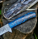 A catfish with a hook in its mouth lies beside the Texan Knives Custom Handmade Damascus Fillet Knife – 13" Pine Cone Handle with Leather Sheath, displayed on a tree stump.