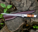 The Texan Knives Custom Handmade Damascus Filleting Knife – 13" with a camel bone handle and leather sheath rests on a weathered stump among green plants.