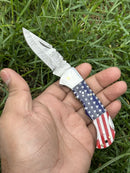 A hand holds the Lone Star 3-Inch Damascus Pocket Knife by Texan Knives, featuring a Damascus steel blade and custom handmade bone handle, against green grass.