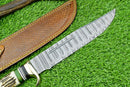 The Texan Knives Deluxe Dundee Knife features a 16" handmade Damascus steel blade, brass guard, pakkawood or stag horn handle, and comes with a leather sheath, shown resting on green artificial grass.