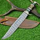 The Texan Knives Custom Handmade Damascus Steel Bowie Knife features a 16" patterned blade, stag horn handle, and comes with a leather sheath, displayed on green grass beside antlers.