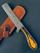The Bull Cutter Knife by Texan Knives features a handmade Damascus steel blade and an orange and black pakkawood handle, displayed on a brown leather sheath with yellow stitching against a dark background.